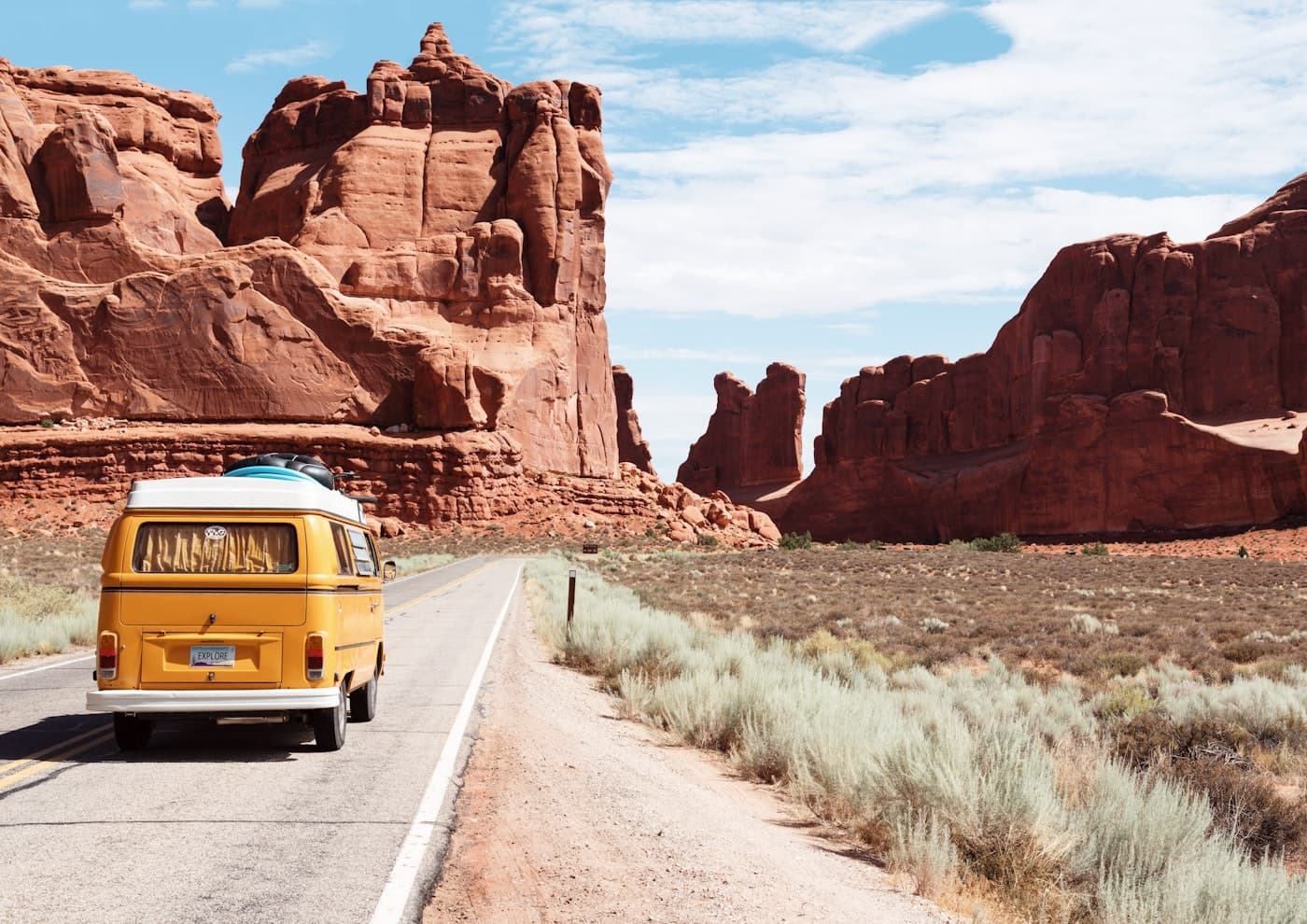 A yellow van on a desert road toward red rock.